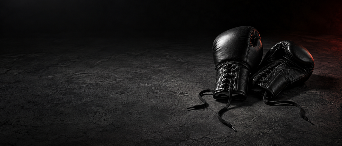 Paire de gants de boxe en cuir noir, lacets défaits, posés sur un sol en béton sombre