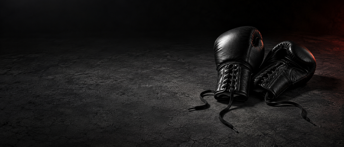 Paire de gants de boxe en cuir noir, lacets défaits, posés sur un sol en béton sombre