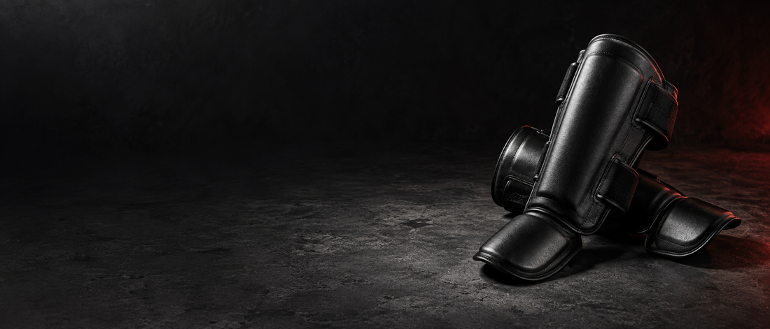 Paire de protège-tibias de boxe thaï noirs à sangles velcro, posés sur un sol en béton sombre