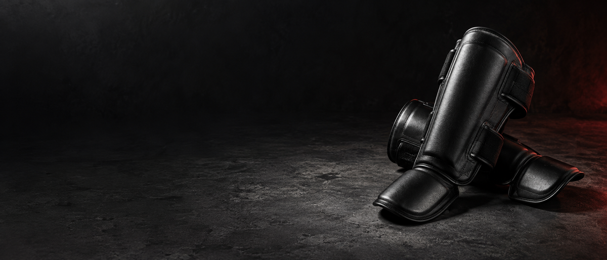 Paire de protège-tibias de boxe thaï noirs à sangles velcro, posés sur un sol en béton sombre